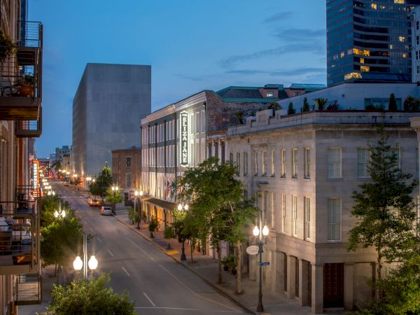 A quiet city street at dusk with historic buildings, streetlamps glowing, and trees lining the sidewalks.