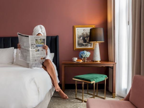 A person sits on a hotel bed reading a newspaper, with a bedside table, lamp, plant, and a pink armchair in the room, bright natural light.