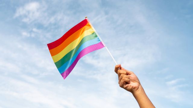 A hand raises a small rainbow Pride flag against a blue sky with light clouds, symbolizing LGBTQ pride.