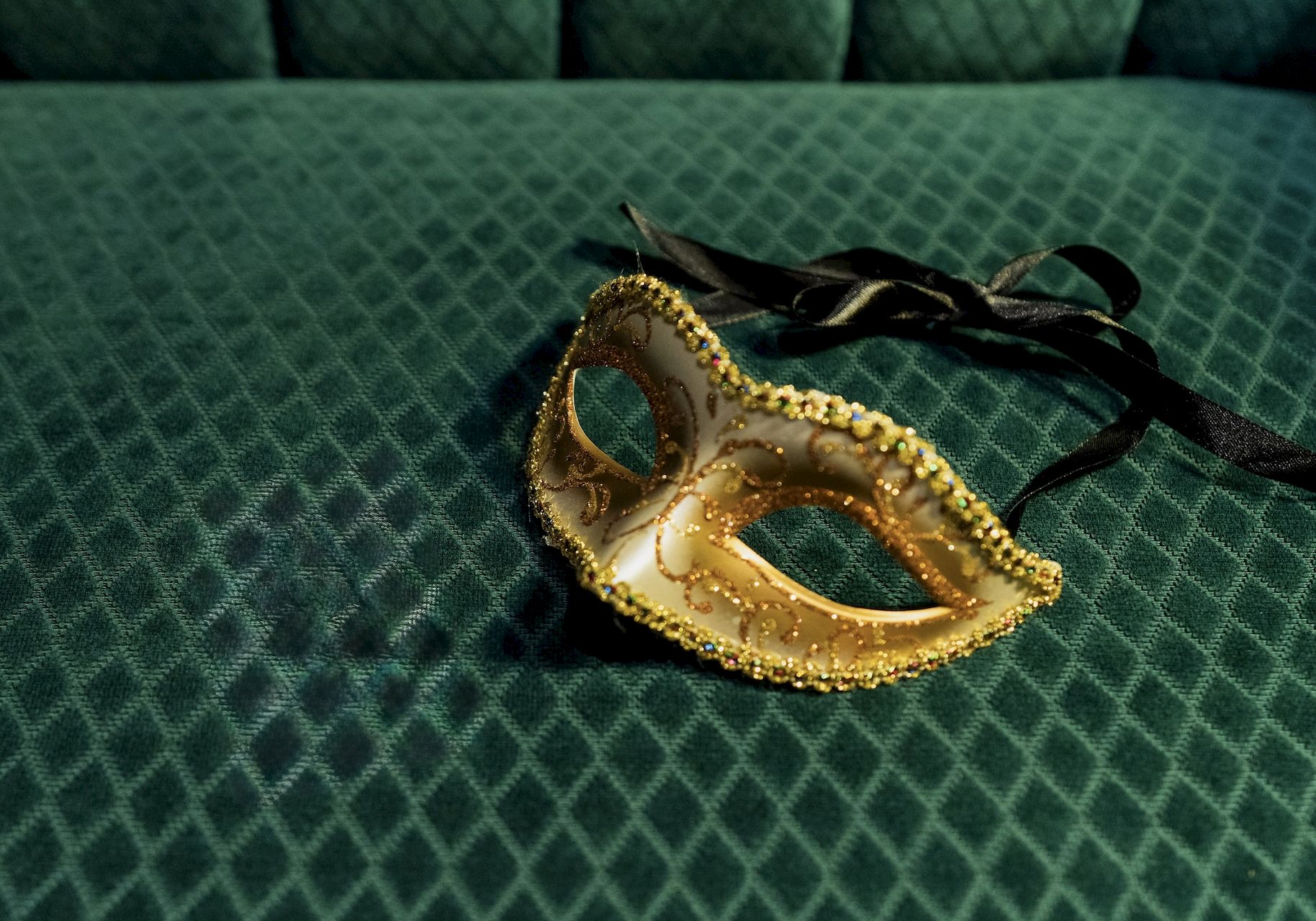 A gold decorative masquerade mask with black ribbon, resting on a green quilted surface.