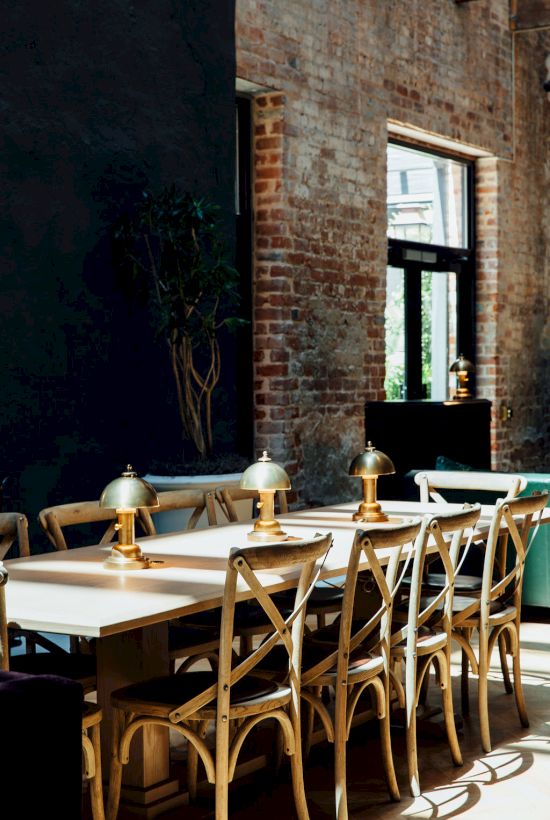 A stylish dining area with a long wooden table, matching chairs, industrial brick walls, and brass desk lamps along the center, bright natural light enters through a window, creating a warm, modern cafe vibe.