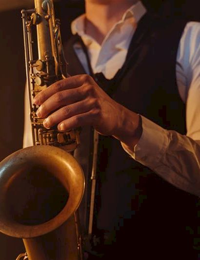 A person playing a saxophone, wearing a white shirt and vest, in a warm-lit setting.
