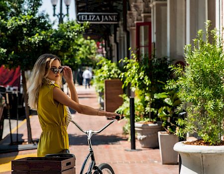 A woman in a yellow outfit rides a vintage bicycle on a sunny sidewalk lined with potted plants and stylish shops, wearing sunglasses.