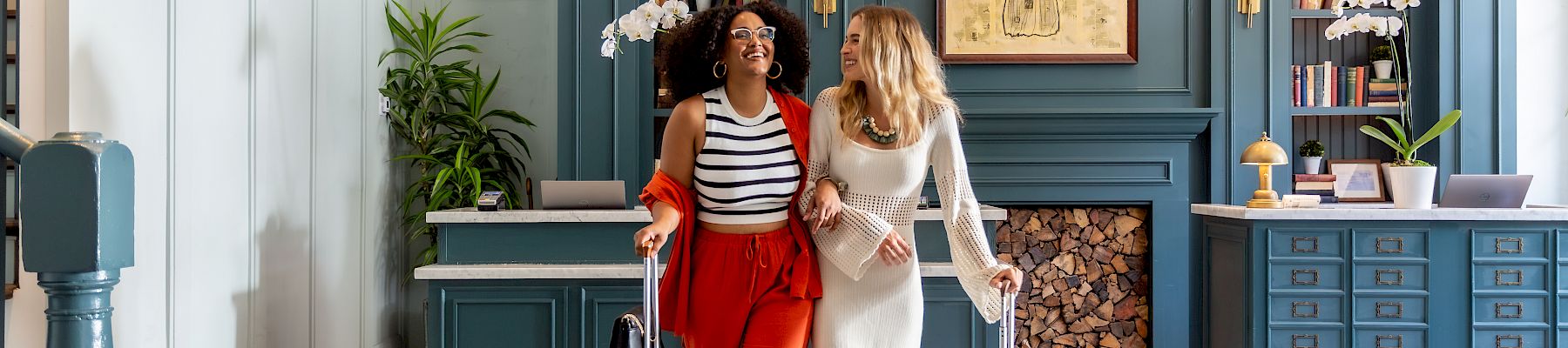 Two women with luggage stand in a stylish hotel lobby, smiling and chatting as they prepare to check in.