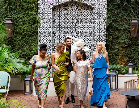 A group of stylishly dressed people walk together down a brick path, smiling and chatting near a patterned archway in a lush garden.