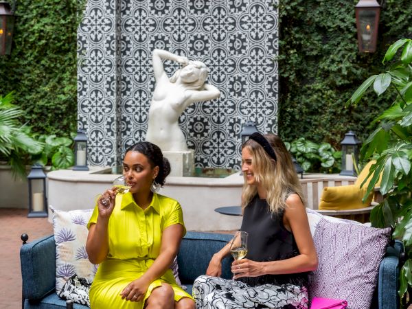 Two stylish women chat on a bench beneath a tiled arch, a white dog sits regally behind them, while snacks and drinks await on the table.