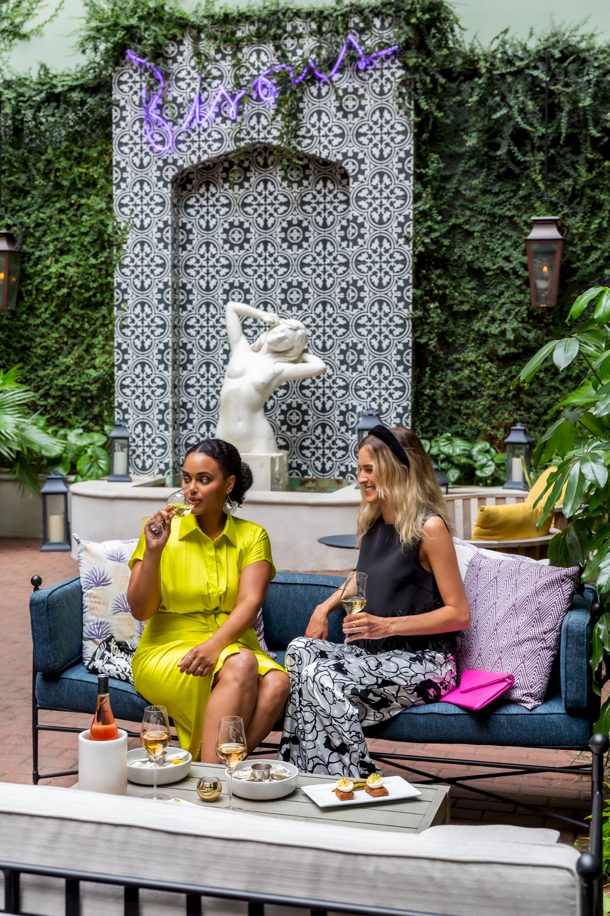 Two stylish women dine outdoors on a terrace, chatting in bright outfits as a white statue dog looks on, amid plants, cushions, and desserts.