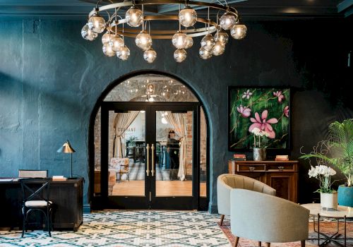 A stylish lounge interior with a hanging chandelier, open doorway to a restaurant, a floral wall art, and cozy seating, lit warmly.