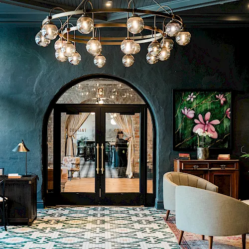 A stylish lounge interior with a hanging chandelier, open doorway to a restaurant, a floral wall art, and cozy seating, lit warmly.