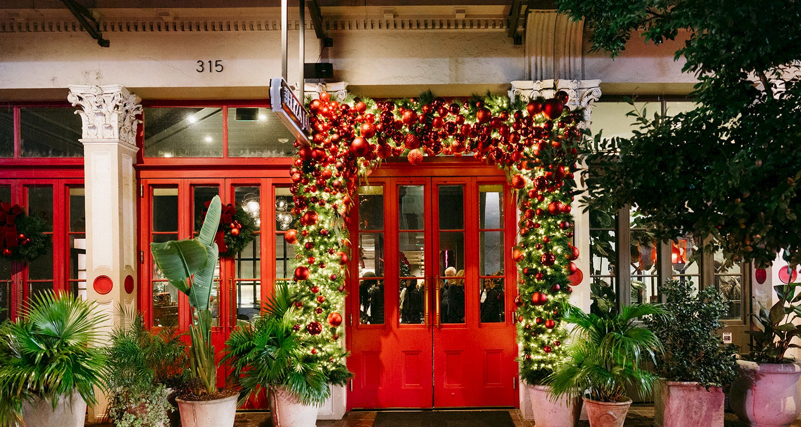 A festive doorway: red double doors covered in lush Christmas garlands and shiny red ornaments, surrounded by potted plants on a patio.