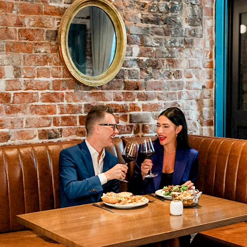 A couple in a restaurant clinks wine glasses and smiles while dining against a brick wall.
