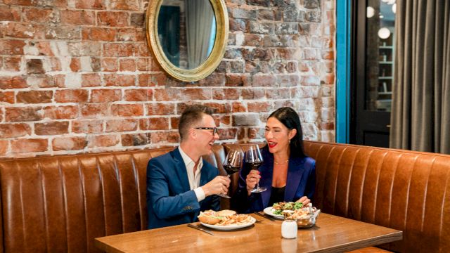 A couple in a restaurant clinks wine glasses and smiles while dining against a brick wall.